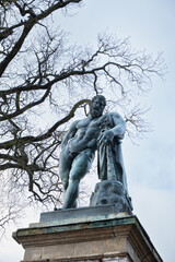 A statue of Hercules depicts a muscular male figure clutching a staff, surrounded by leafless trees and a cloudy sky in a public park in the city of Pushkin, Russia - March 04, 2025