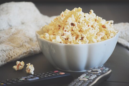 Close-up of a bowl of popcorn and a knitted jumper on a  table next to two TV remote controls
