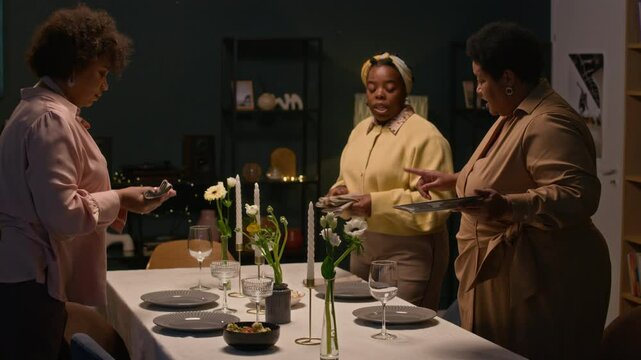 Medium full shot of jolly Black woman dressed in elegant outfit serving dinnerware on festive dining table while lively chattering with elder family members assisting in setting home celebration