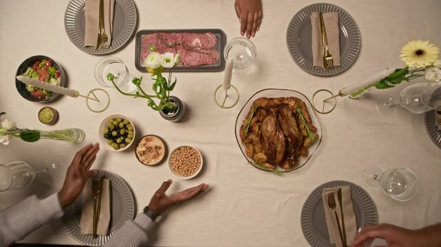 Flat lay shot of family members serving roasted turkey with appetizers on dining table decorated with candles and white tablecloth at festive dinner