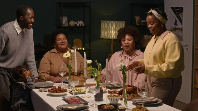 Cheerful Black woman dressed in elegant outfit serving plate of roasted chicken on dining table then discussing festive dish recipe with family members during cozy home gathering