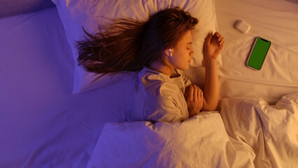 Young woman sleeping in bed, laying head on the pillow covered with blanket in purple neon light, wearing pods, smartphone with chroma key green screen background on the bed. Top view