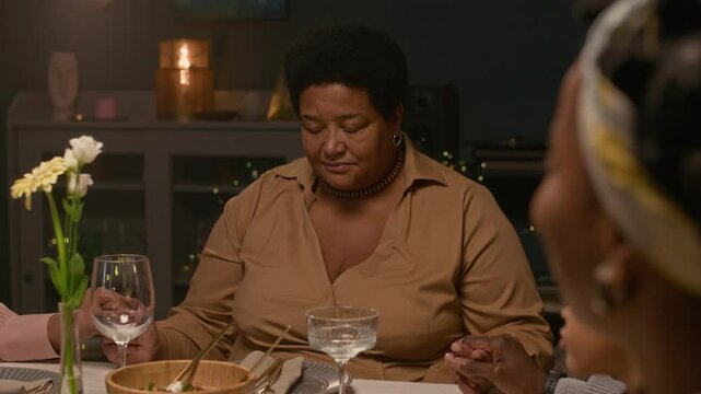 Medium shot of Black granny saying grace before festive home dinner sharing meal with adult children and whole extended family while sitting together around dining table, rack focus