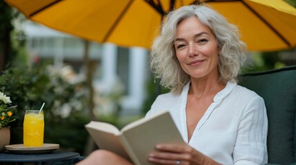 A woman reading a book while sitting under an umbrella, enjoying a glass of orange juice, outdoors on a sunny day.