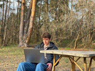 Obraz premium a young man is sitting in a park and working online on his laptop, the concept of remote work from anywhere in the world