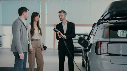 Caucasian husband and wife standing next to sales manager at car dealership. Male explaining...