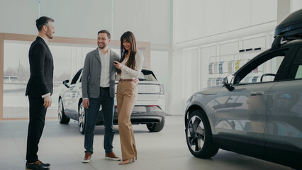 Caucasian couple standing next to new purchased car in showroom. Attractive dealer in elegant suit...