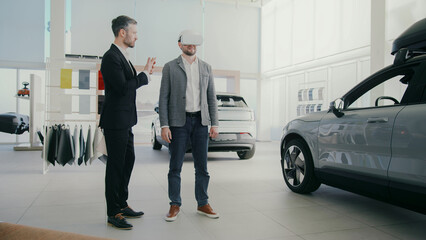 Man using VR headset while standing in modern car dealership. Professional dealer in black suit standing next to customer and telling something. Exploring virtual car or product. Technology concept.