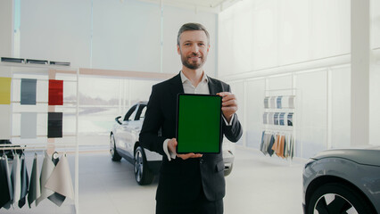 Fototapeta premium Handsome Caucasian man standing in modern showroom. Holding tablet device with chroma key. Manager wearing suit and smiling warmly. Modern auto salon for selecting and buying vehicles.