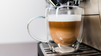 Home coffee maker pouring cappuccino into glass mug in kitchen