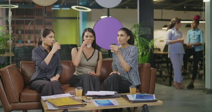 Indian asian young adult gen z girl woman sitting on sofa drink coffee give high five indoor job office happy female intern staff talk funny gossip spend break time enjoy fun joy day at work place