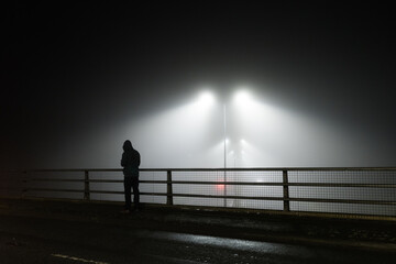 A mysterious unknown hooded figure standing on a bridge over a motoway, silhouetted by street lights on a foggy eerie night