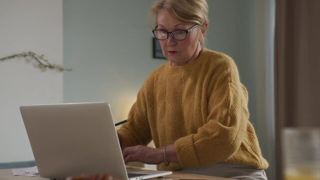 Senior elegant woman sits at table viewing papers and utility bills. Aged woman works remotely using laptop computer. Older focused businesswoman making notes. Female elegant writer creative process