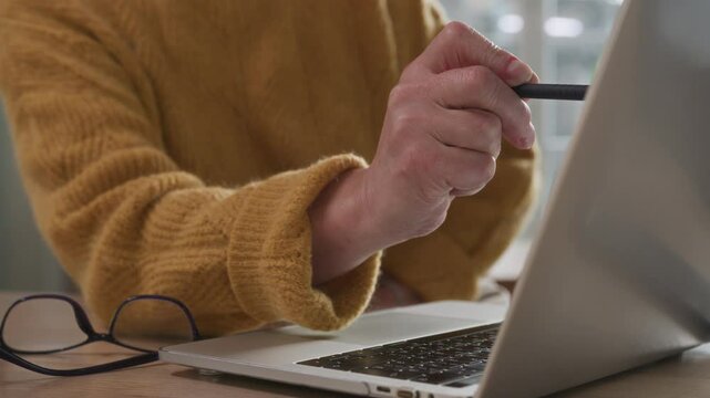 Senior elegant woman sits at table viewing papers and utility bills. Aged woman works remotely using laptop computer. Older focused businesswoman making notes. Female elegant writer creative process