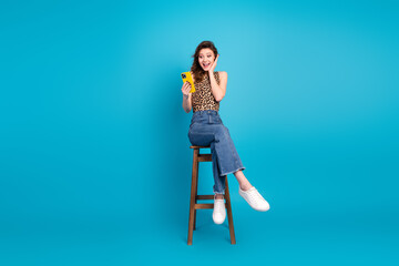 Young woman sitting on a stool, holding a yellow smartphone, smiling surprised against a solid blue background.