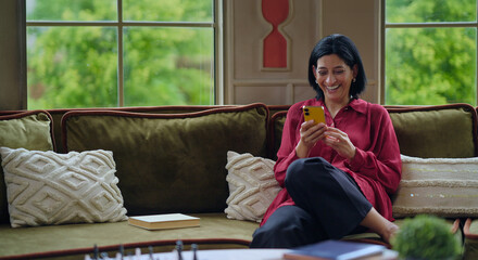 Indian happy relax gen x woman lady sitting sitting on cozy sofa couch hold mobile typing text sms...