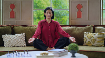 Indian happy gen x relax mind yogini woman lady mom cross leg sitting cozy sofa couch closed eyes do health care well being yoga asana take deep breath fresh air feel good enjoy day time indoor home