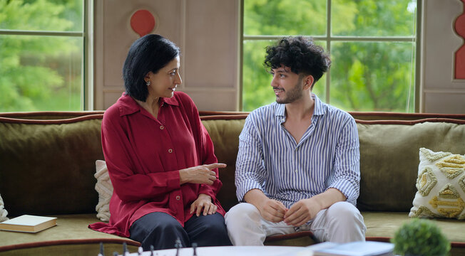 Indian happy mature woman lady with young man male sitting on cozy sofa couch talk gossip share story indoor home adult mom teen son boy speak funny joke enjoy day spend time have fun joy inside house