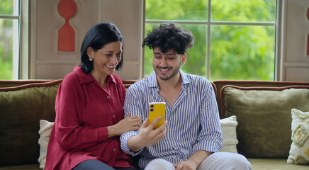 Indian Asian happy older woman mom lady young adult man male son sitting cozy sofa couch use mobile phone talk video call make funny gossip look cam screen joy enjoy fun day spend time indoor house