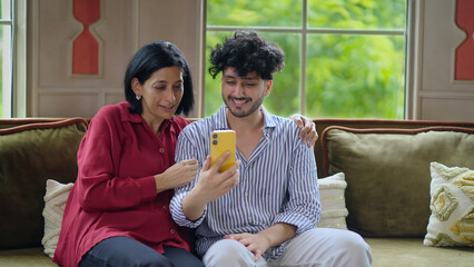 Indian Asian happy older woman mom lady young adult man male son sitting cozy sofa couch use mobile phone talk video call make funny gossip look cam screen joy enjoy fun day spend time indoor house