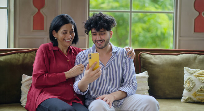 Indian Asian happy older woman mom lady young adult man male son sitting cozy sofa couch use mobile phone talk video call make funny gossip look camera joy enjoy fun day spend time indoor house