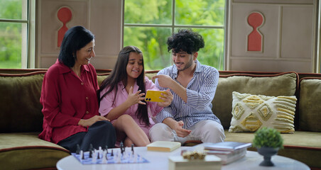 Indian mom teen girl boy sitting on cozy sofa couch hold mobile phone look cam talk funny gossip enjoy online video call indoor home happy woman lady young adult man male have fun joy spend day time