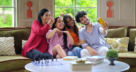 Indian Asian happy mom lady with young adult child sitting cozy sofa couch use mobile take selfie indoor home cute teen girl boy hold phone look cam make funny pose have fun joy enjoy day spend time