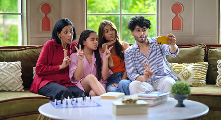 Indian Asian happy mom lady with young adult child sitting cozy sofa couch use mobile take selfie indoor home cute teen girl boy hold phone look cam make pout pose have fun joy enjoy day spend time