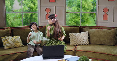 Indian happy cute child kid with young gen z girl sitting on cozy sofa couch use laptop listen online mp3 music teen boy adult woman lady sing song spend time enjoy day have fun joy indoor home house