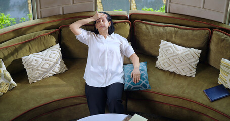Indian gen x alone woman lady sitting on sofa couch have health issues touch head indoor home upset...
