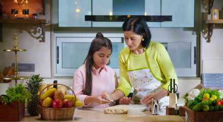 Indian happy woman lady with cute teen age cute girl rolling raw flour dough learn cooking food indoor home smiling young mom teach to kid child baking cookies enjoy day indoor modern home kitchen