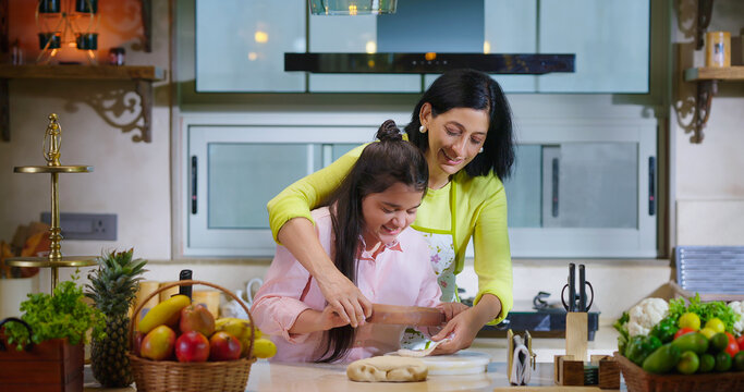 Indian happy woman lady with cute teen age cute girl rolling raw flour dough learn cooking food indoor home smiling young mom teach to kid child baking cookies enjoy day indoor modern home kitchen