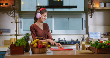 Indian Asian happy young adult gen z woman lady wear headphone online music mp3 enjoy day sing song spend time fun joy cute teen girl female hold knife cut tomato slice on chop board indoor kitchen