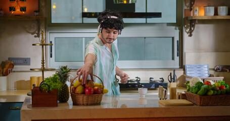 Indian happy young adult gen z man guy listen online mp3 song music have fun joy indoor kitchen teen boy male take fresh apple fruits from basket enjoy day spend time