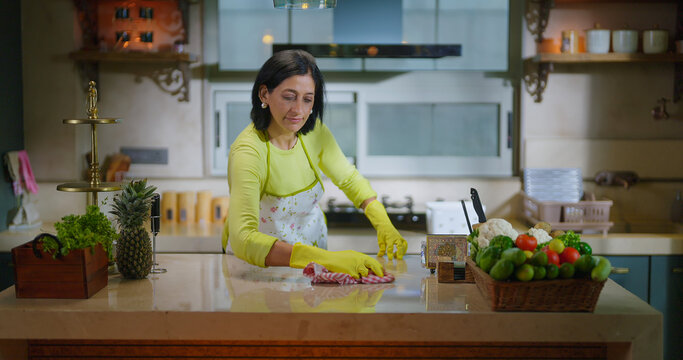 Indian gen x mature adult mom wear apron and hand gloves collect pick up waste trash in box use rag. Middle aged woman lady do work use rag wiping dust on table keep modern kitchen neat clean tidy