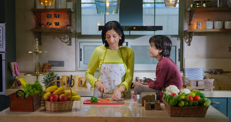 Indian happy mature mom standing indoor modern home kitchen hold knife cut fresh slice on chop board young adult woman lady talk gossip son child boy eat vegan piece enjoy day spend time have fun joy