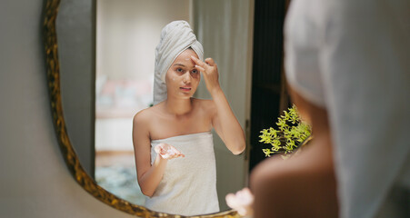 Indian young adult gen z woman lady wrapped towel on head and body look mirror apply make up cream on dry cheek face teen age girl female hold bottle take liquid hand do fresh day skin care house home