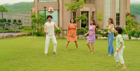 Indian happy family young man woman group play ball game throwing in air have fun joy enjoy outdoor park garden Playful cute teen girl son child kid boy with adult mom lady dad spend day time outside	