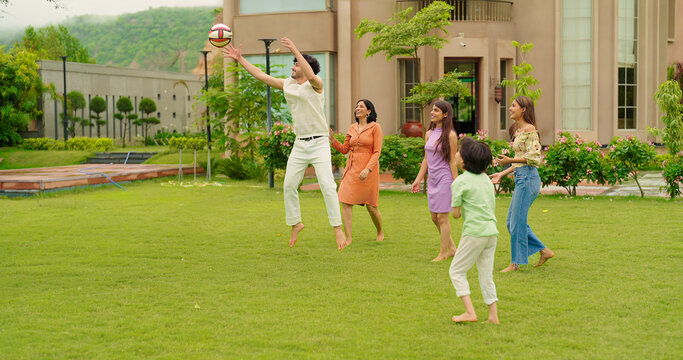 Indian happy family young man woman group play ball game throwing in air have fun joy enjoy outdoor park garden Playful cute teen girl son child kid boy with adult mom lady dad spend day time outside	