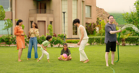 Indian happy dad man hold hose pipe spray water on family group outdoor house lawn park young adult teen girl mom cute son kid boy child male female play game have fun joy spend time enjoy picnic trip