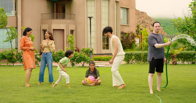 Indian happy dad man hold hose pipe spray water on family group outdoor house lawn park young adult teen girl mom cute son kid boy child male female play game have fun joy spend time enjoy picnic trip - Powered by Adobe