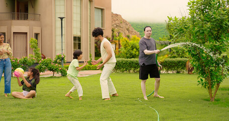 Indian happy dad man hold hose pipe spray water on family group outdoor house lawn park young adult...
