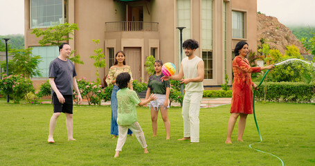 Indian happy mom hold hose pipe spray water on house lawn park tree young adult family teen girl dad cute son kid boy child male man female group play ball game have fun joy spend day time enjoy trip 