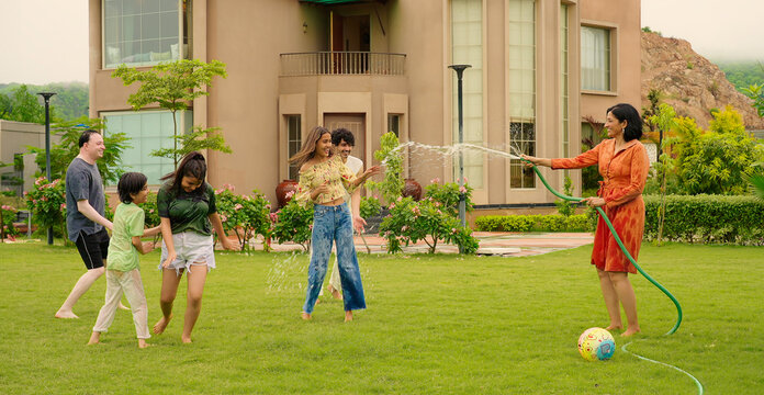 Indian happy mom hold hose pipe spray water on young adult family teen girl dad cute son kid boy child male man female group play ball game have fun joy spend time enjoy day trip house lawn park - Powered by Adobe