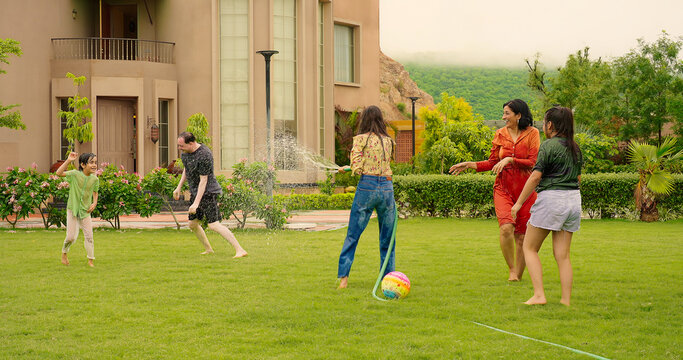 Indian happy teen girl lady mom hold hose pipe spray water on family group play game house lawn cute kid boy son child young adult man have fun joy day spend funny time enjoy trip outside picnic park
