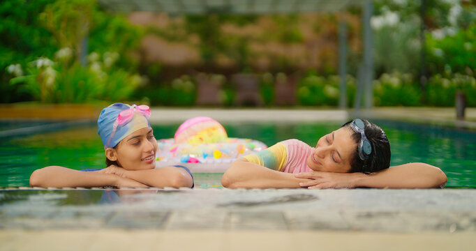 Indian relax wet adult mom woman with cute teen girl standing take rest on pool edge feel tired. Two young female talk gossip at outdoor water park resort trip spend time luxury hotel villa enjoy day