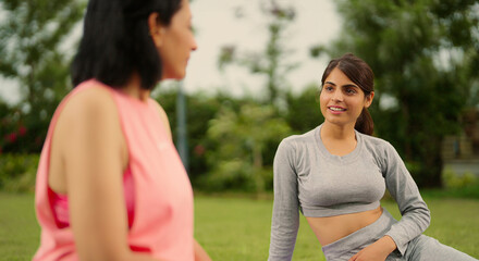 Fototapeta premium Indian relax woman lady mom teen age girl sitting on mat talk gossip about self body health care good habit. Two happy young adult female take break after warm up spend day time outdoor garden park