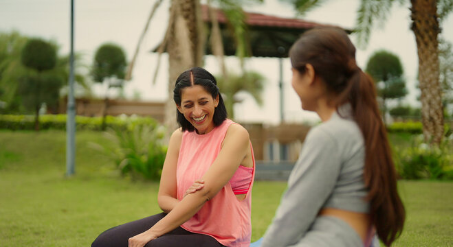 Indian relax woman lady mom teen age girl sitting on mat talk gossip about self body health care good habit. Two happy young adult female take break after warm up spend day time outdoor garden park - Powered by Adobe