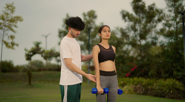 Indian young adult man woman use dumbbells do hand lifting muscle workout outdoor garden park healthy fit boy male guy standing teach to female girl good habit learn gym warm up self health body care