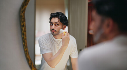 Indian Asian gen z young adult smiling guy bearded man look mirror hold soft brush apply white foam gel cream to his beard face before shave do morning day self skin care routine indoor house